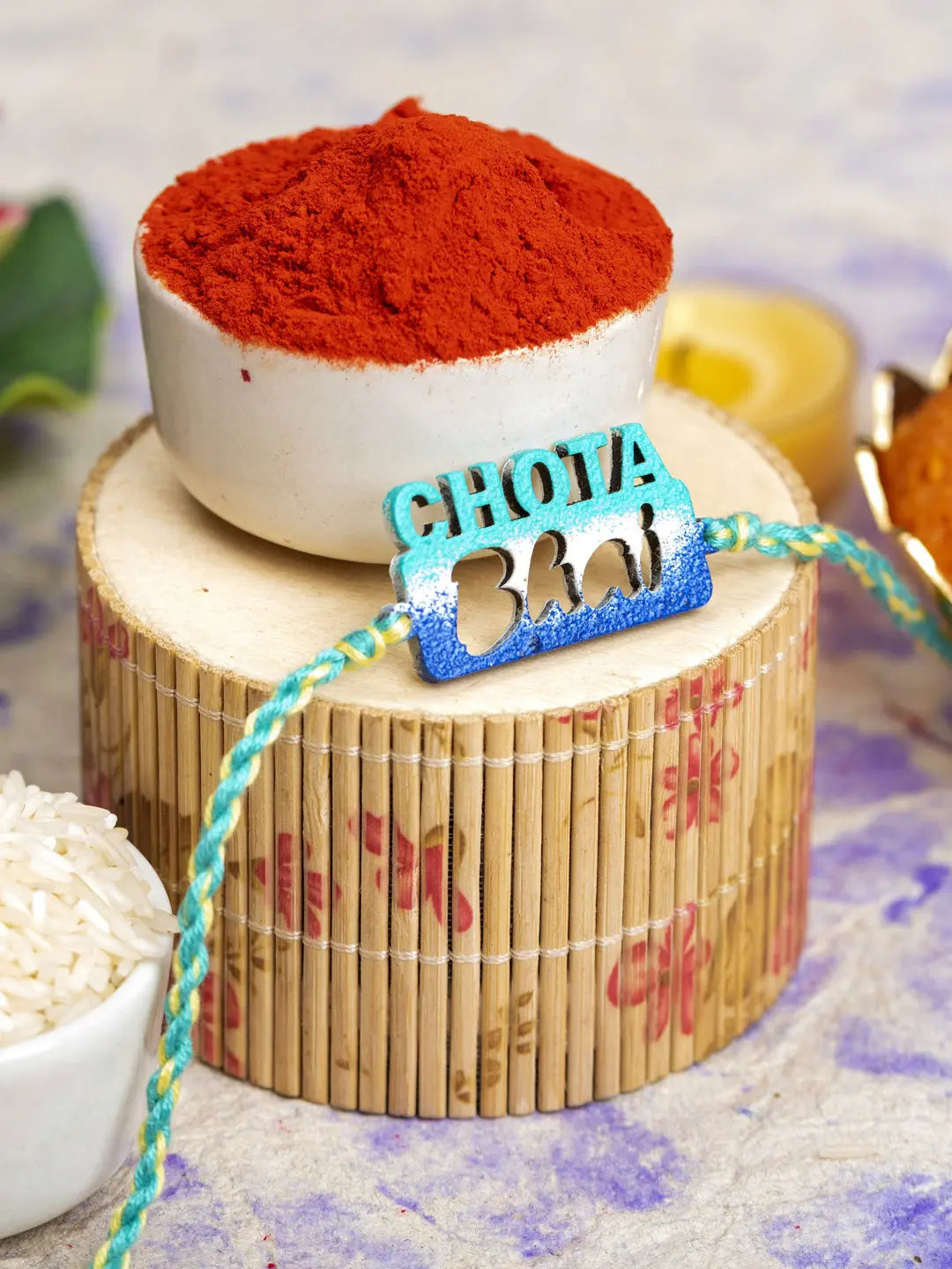 LOOK AND ADORN chhota bhai rakhi with blue and green thread placed on wooden decor with red powder in background