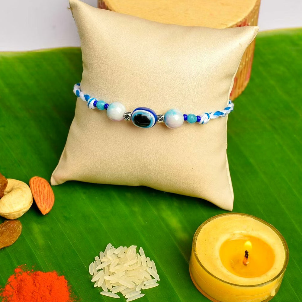 LOOK AND ADORN evil eye rakhi with blue and white beads displayed on a beige cushion with traditional Indian ritual items