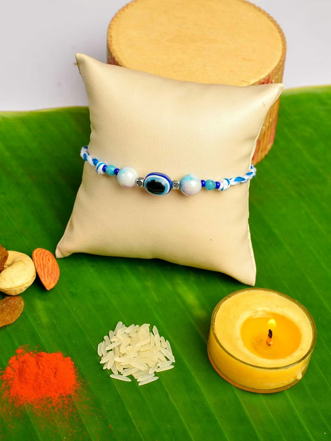 LOOK AND ADORN evil eye rakhi with blue and white beads displayed on a beige cushion with traditional Indian ritual items