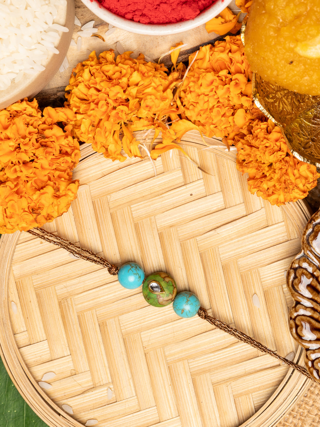 Kundan rakhi with blue and green beads on a traditional bamboo plate surrounded by marigold flowers and festive items