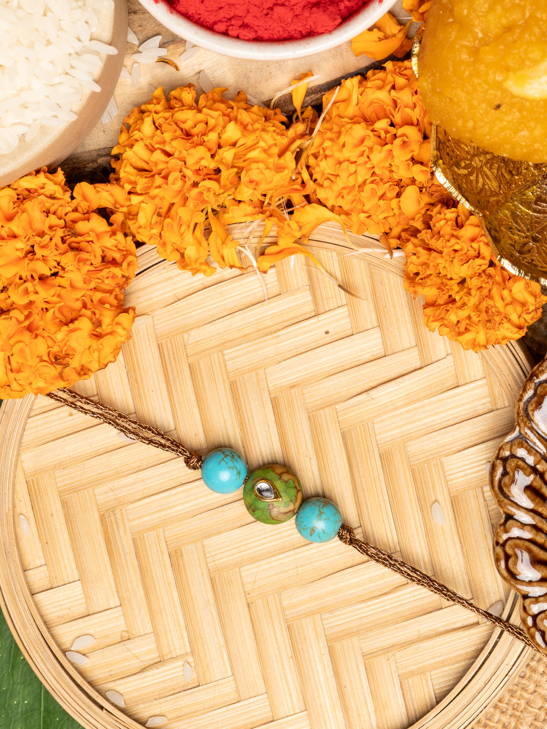 Kundan rakhi with blue and green beads on a traditional bamboo plate surrounded by marigold flowers and festive items