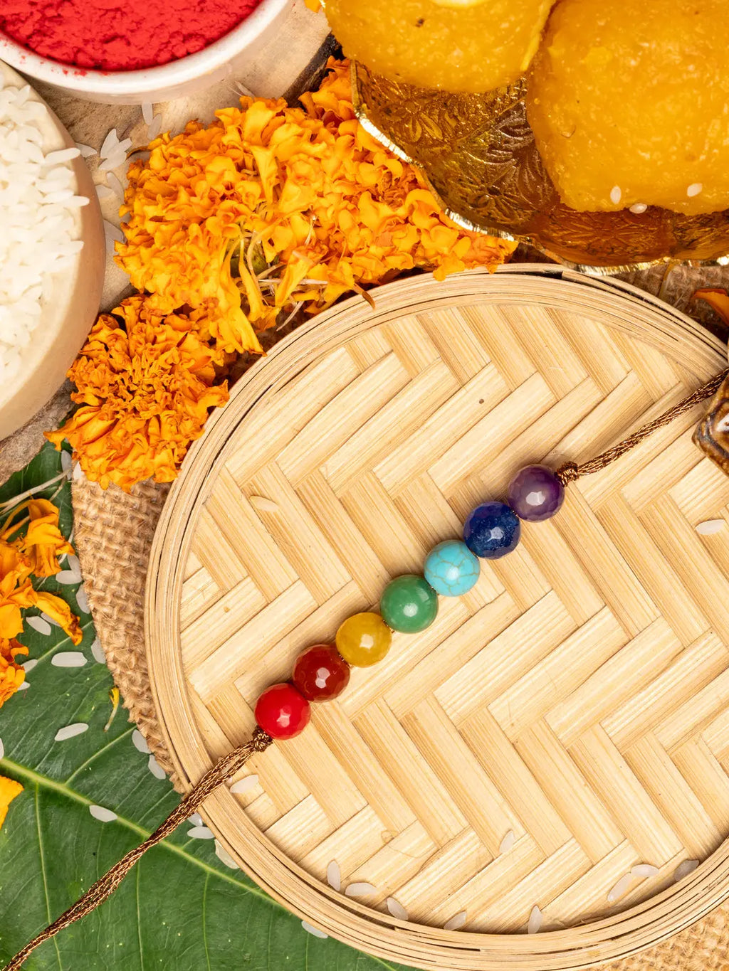 7 chakra rakhi with colorful beads on wooden tray surrounded by marigold flowers and traditional sweets