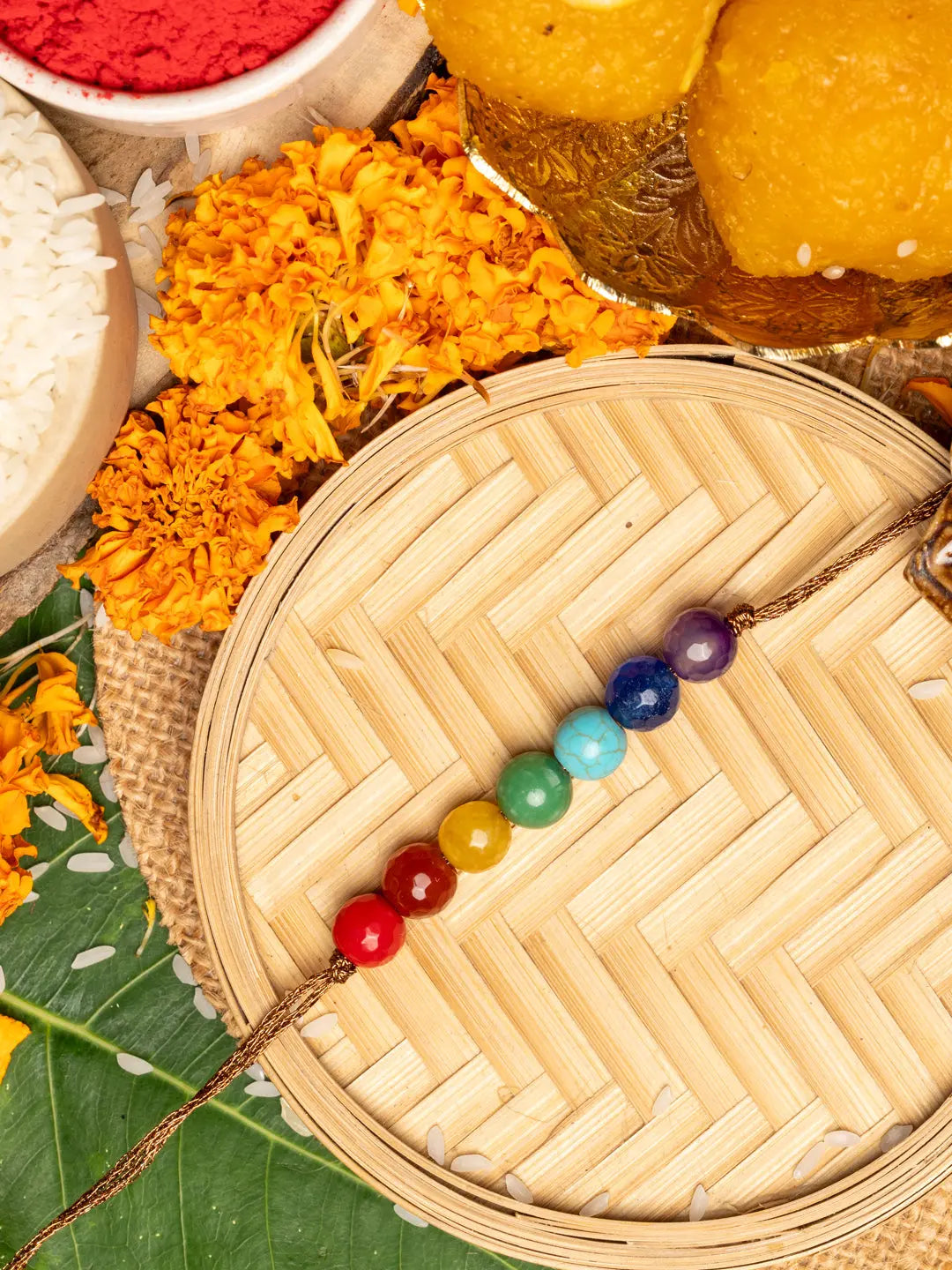 7 chakra rakhi with colorful beads on wooden tray surrounded by marigold flowers and traditional sweets