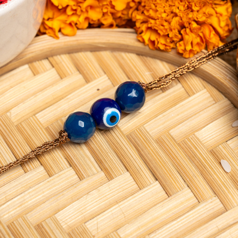 LOOK AND ADORN evil eye rakhi with blue beads on golden thread placed on bamboo mat with marigold flowers