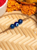 LOOK AND ADORN evil eye rakhi with blue beads on golden thread placed on bamboo mat with marigold flowers