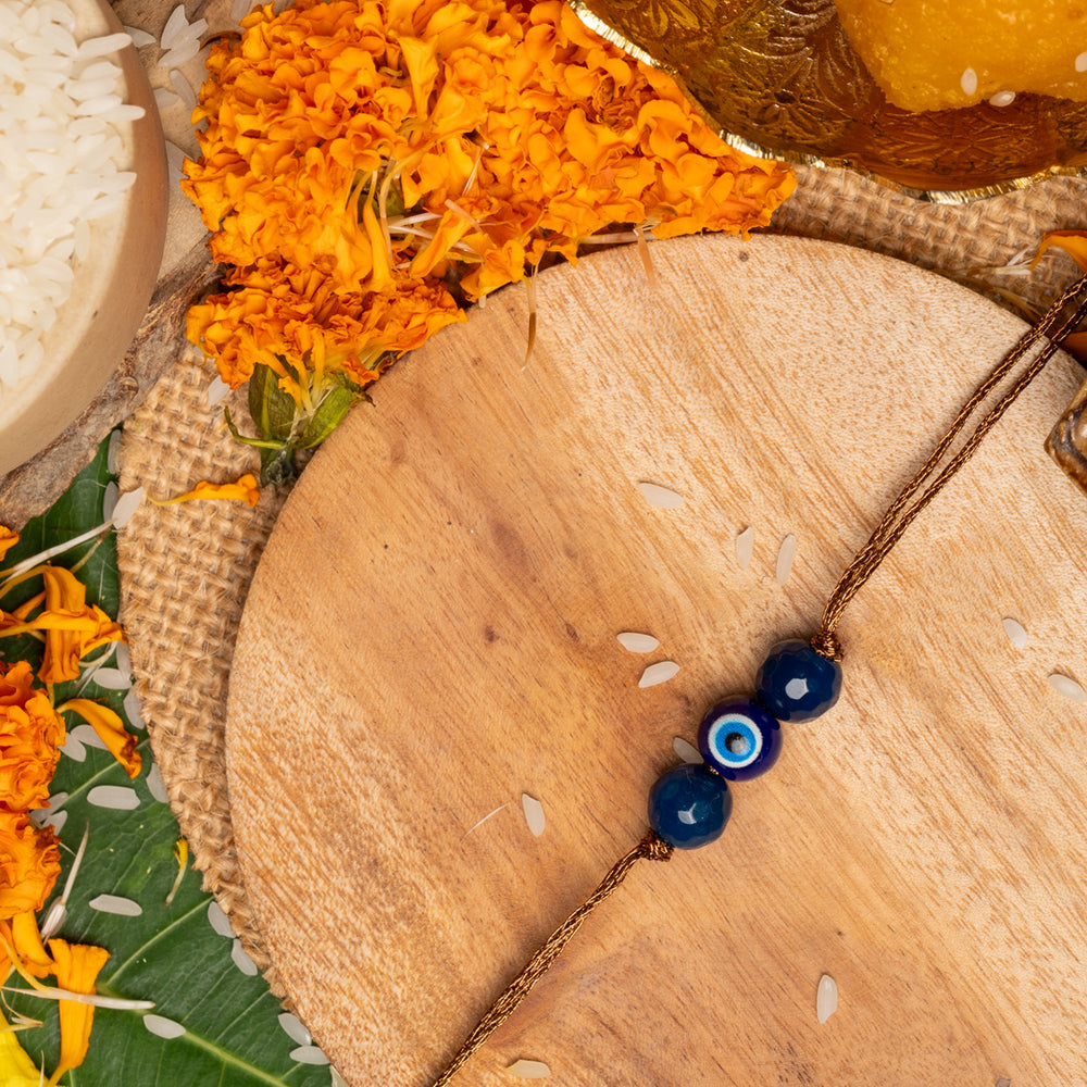 LOOK AND ADORN evil eye rakhi with blue beads on a wooden plate surrounded by marigold flowers and traditional pooja items