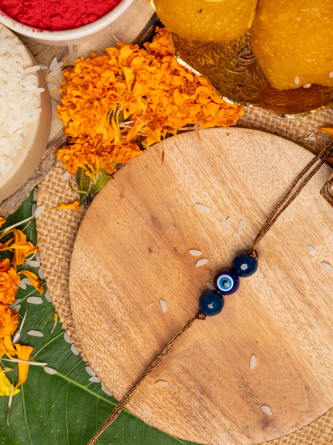 LOOK AND ADORN evil eye rakhi with blue beads on a wooden plate surrounded by marigold flowers and traditional pooja items