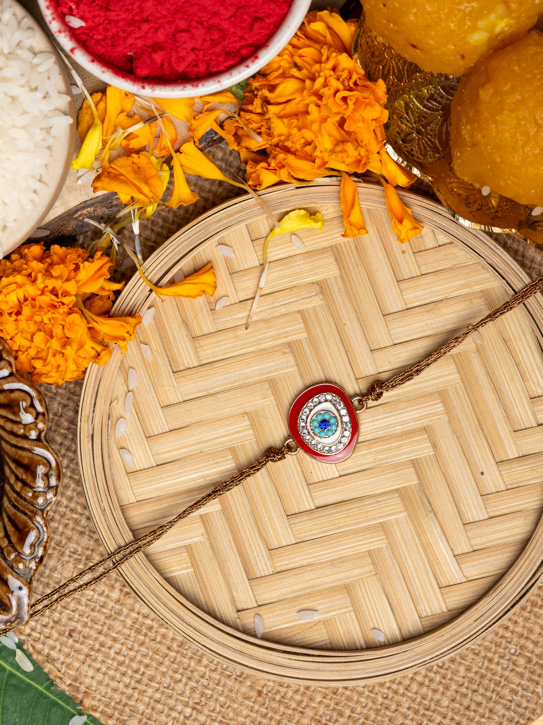 LOOK AND ADORN evil eye rakhi with blue and white beads on a woven tray surrounded by marigold flowers and traditional items