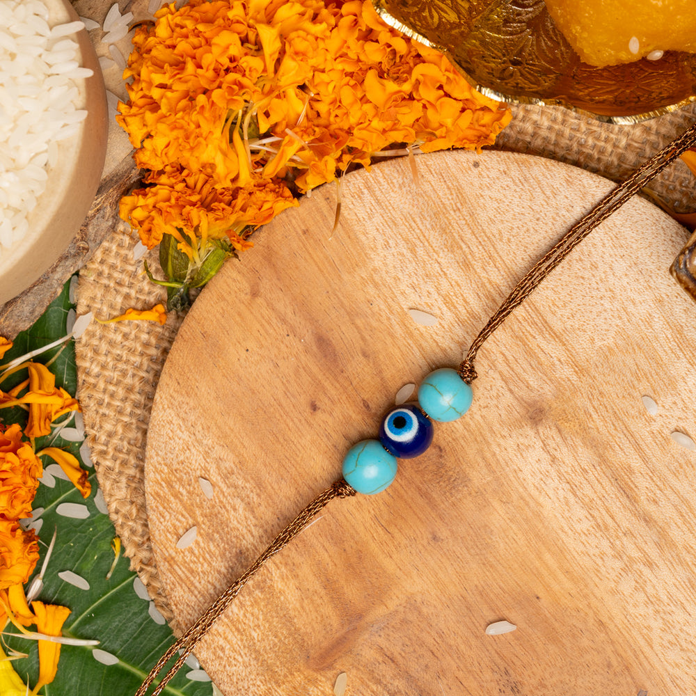LOOK AND ADORN evil eye rakhi with blue beads on wooden plate surrounded by marigold flowers and traditional items