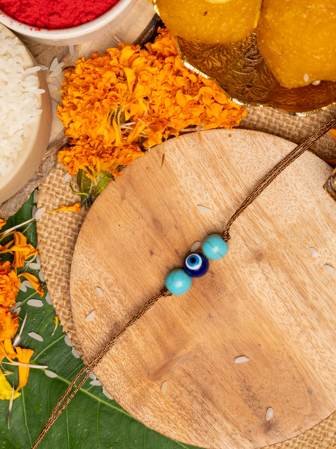 LOOK AND ADORN evil eye rakhi with blue beads on wooden plate surrounded by marigold flowers and traditional items