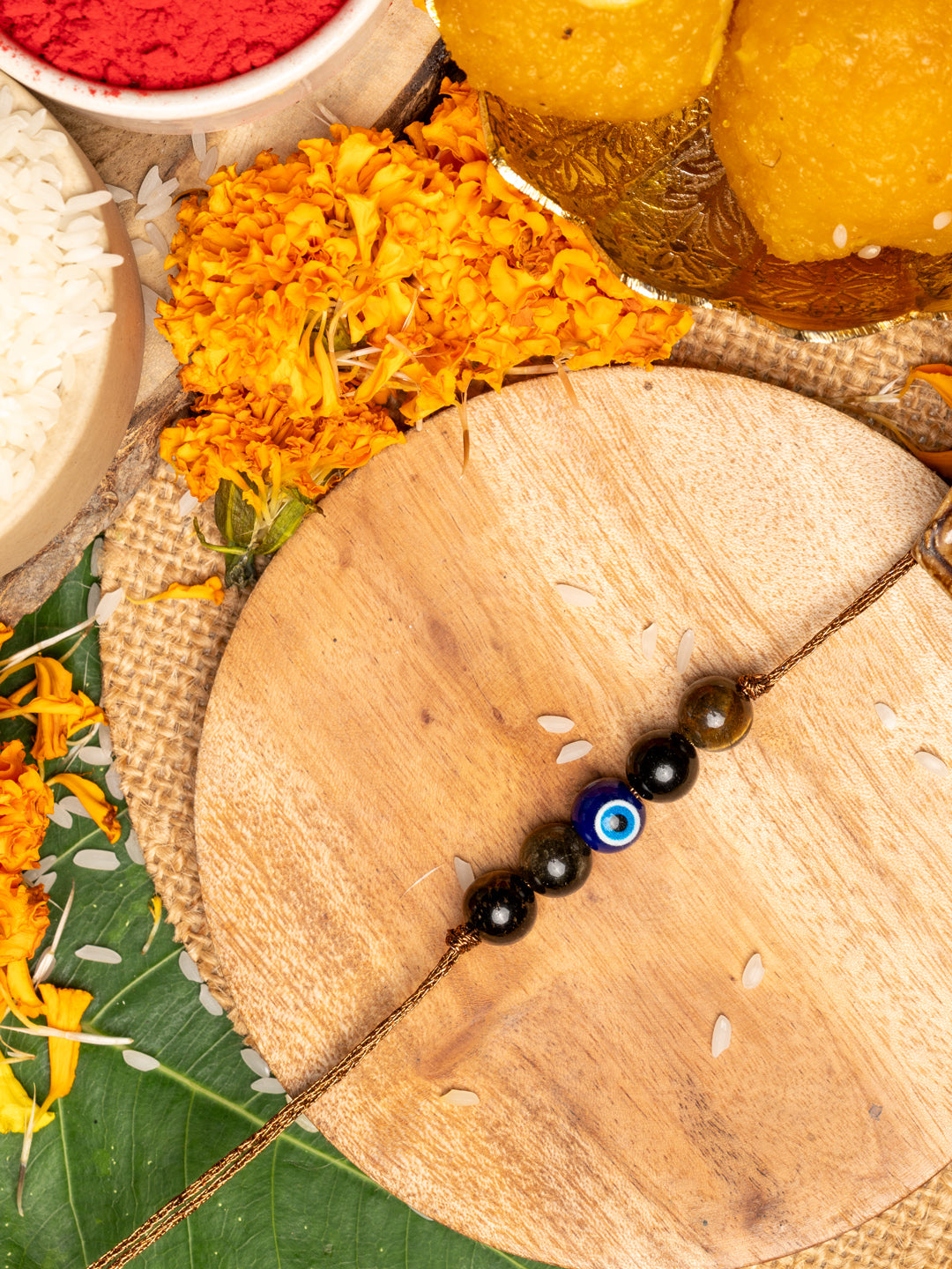 Elegant evil eye rakhi with black beads on wooden plate surrounded by marigold flowers and traditional pooja items