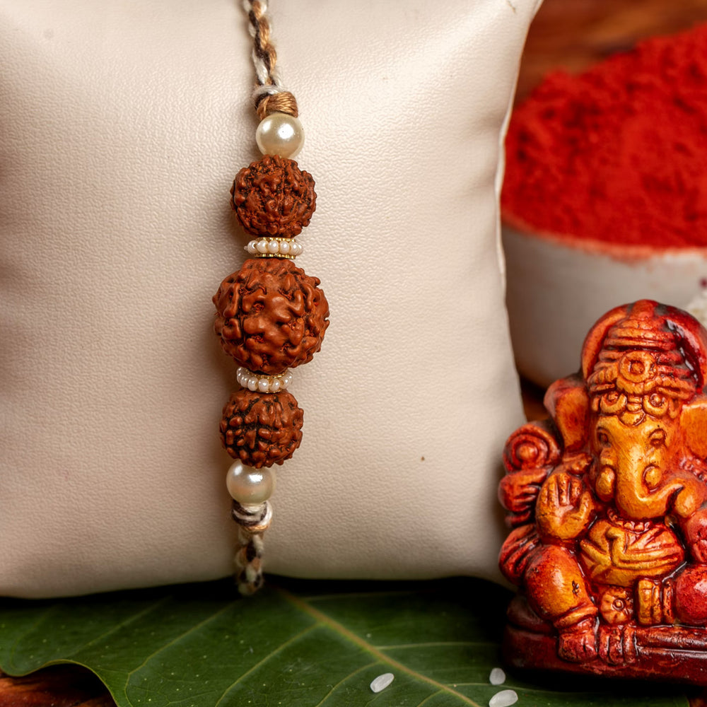 LOOK AND ADORN rudraksh rakhi with three beads and pearl accents displayed on a white cushion next to a small Ganesha idol
