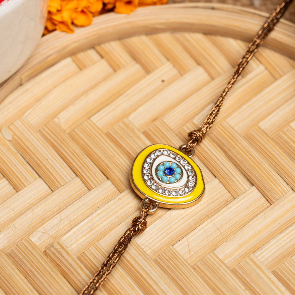 LOOK AND ADORN evil eye rakhi with blue and yellow design and rhinestone embellishments on a woven mat