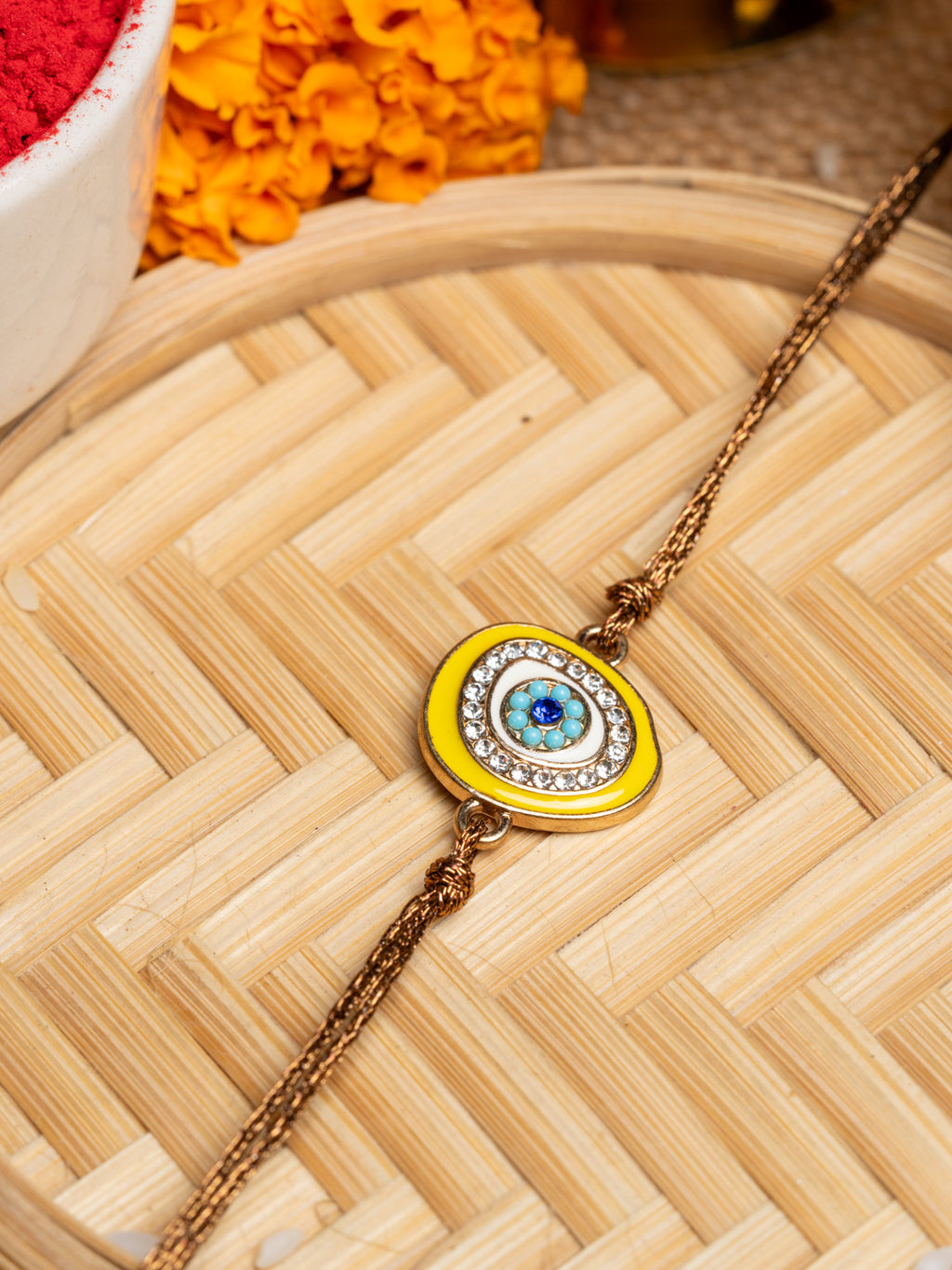 LOOK AND ADORN evil eye rakhi with blue and yellow design and rhinestone embellishments on a woven mat