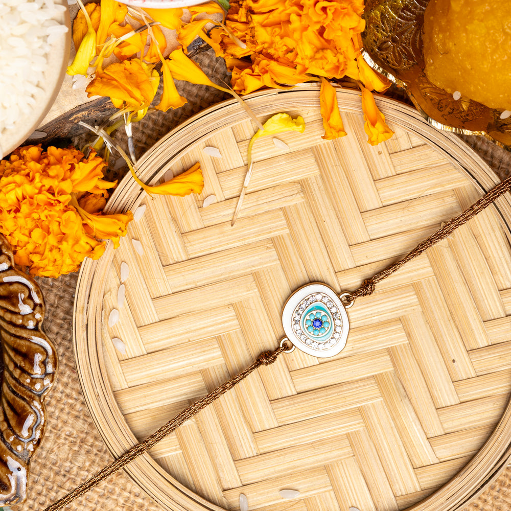 LOOK AND ADORN evil eye rakhi with blue and white charm on a woven tray surrounded by marigold flowers and traditional pooja items