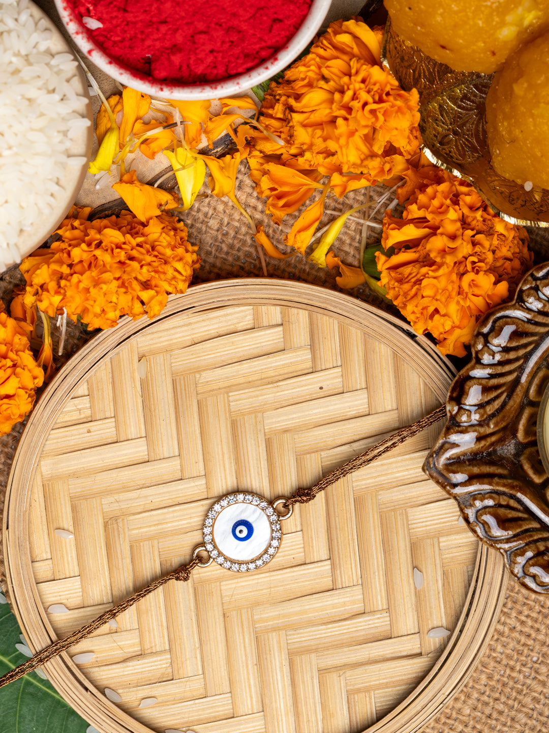 LOOK AND ADORN evil eye rakhi on woven tray surrounded by marigold flowers and traditional puja items