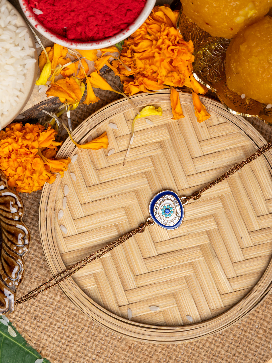 LOOK AND ADORN evil eye rakhi with blue and white charm on a woven plate with marigold flowers and traditional pooja items