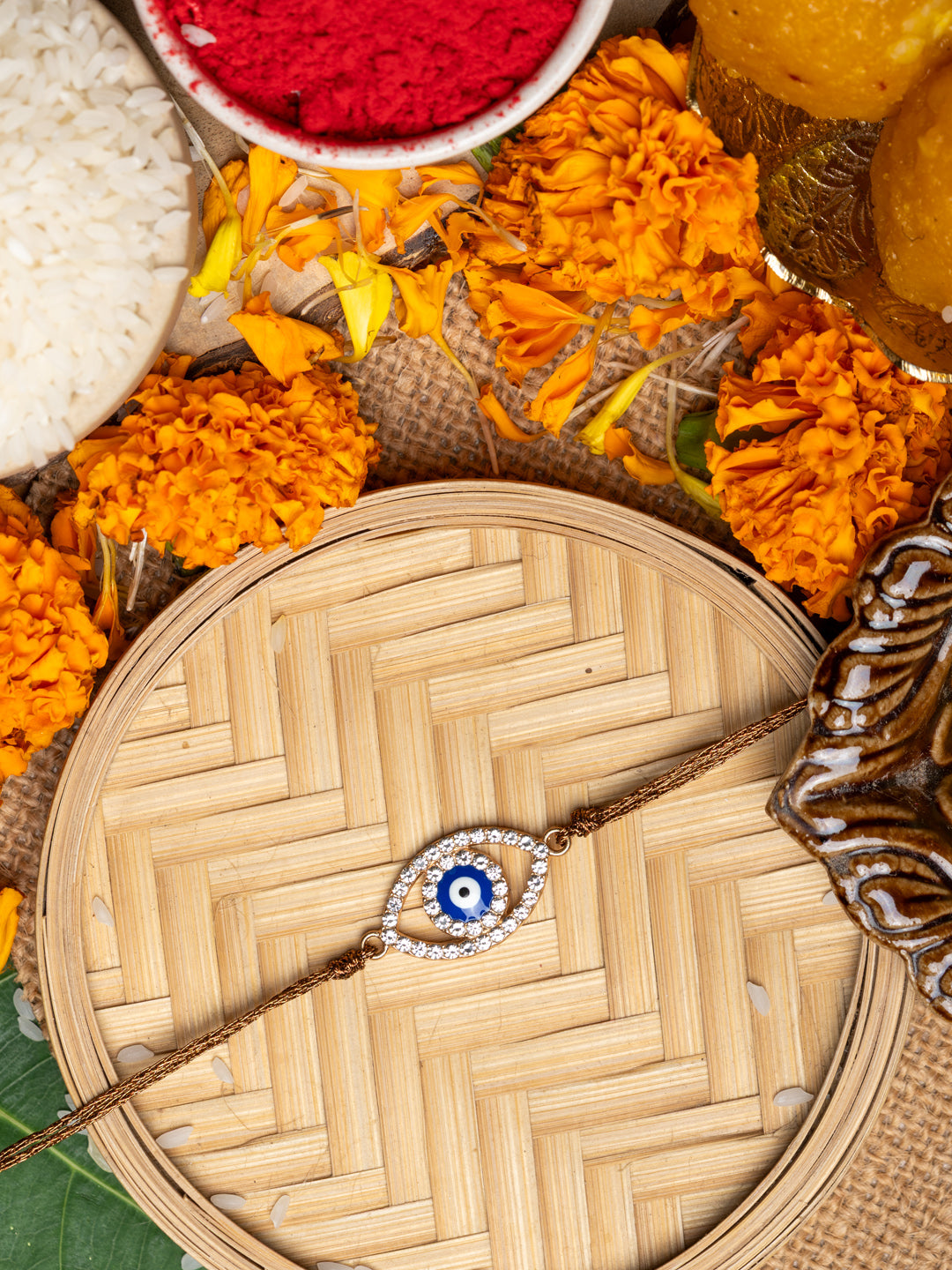 LOOK AND ADORN evil eye rakhi with blue and white bead design surrounded by marigold flowers and traditional pooja items
