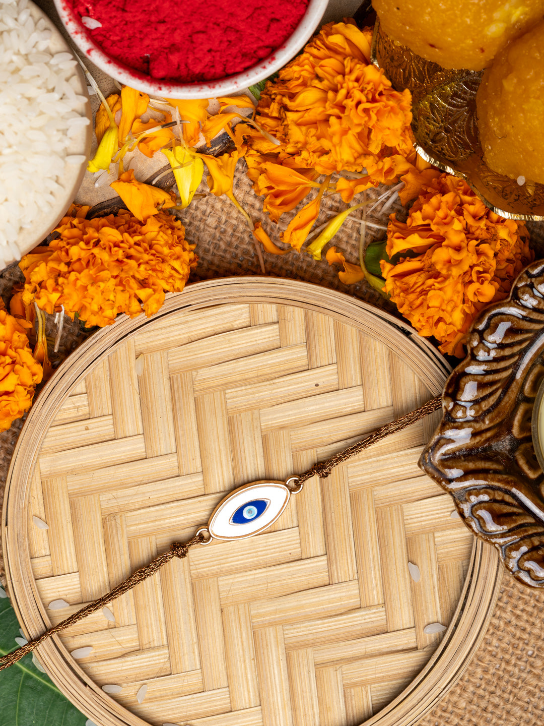LOOK AND ADORN evil eye rakhi with blue eye charm on a woven bamboo plate surrounded by marigold flowers and traditional pooja items