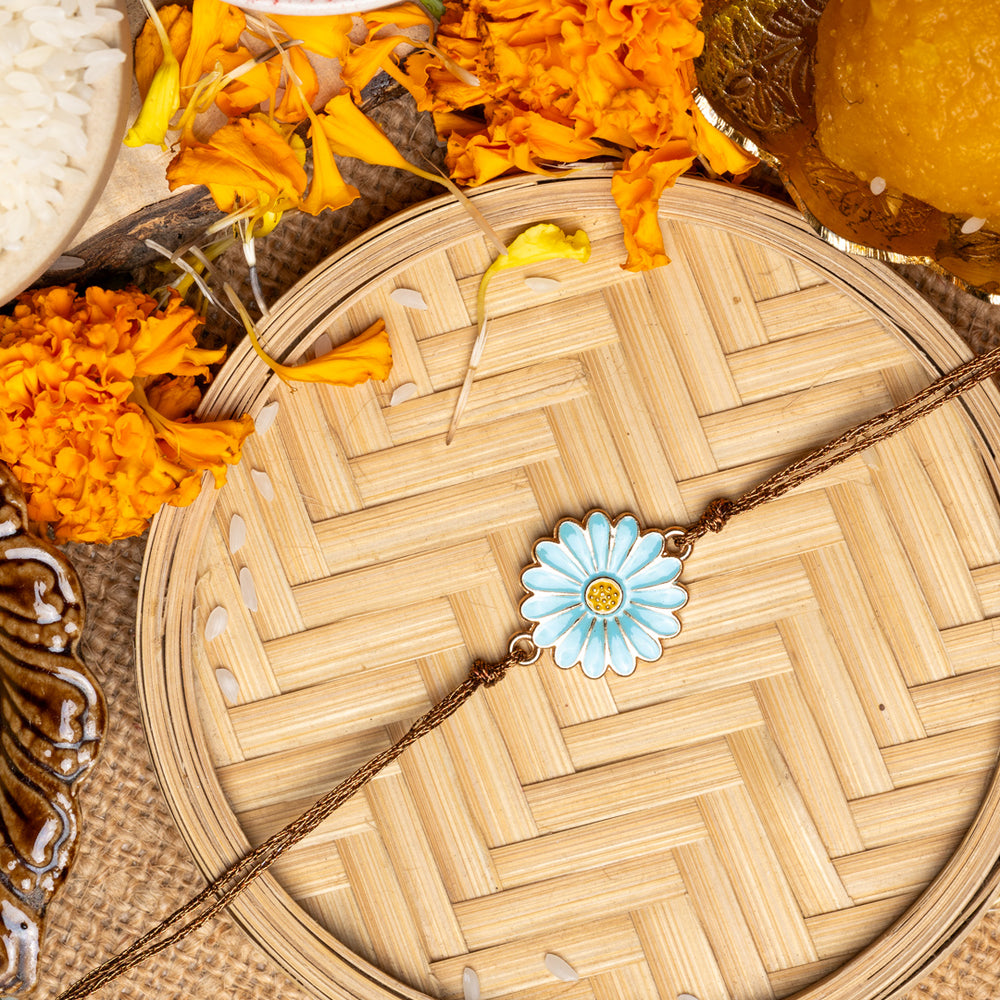 LOOK AND ADORN floral rakhi with blue flower design placed on woven bamboo tray with marigold flowers and traditional sweets