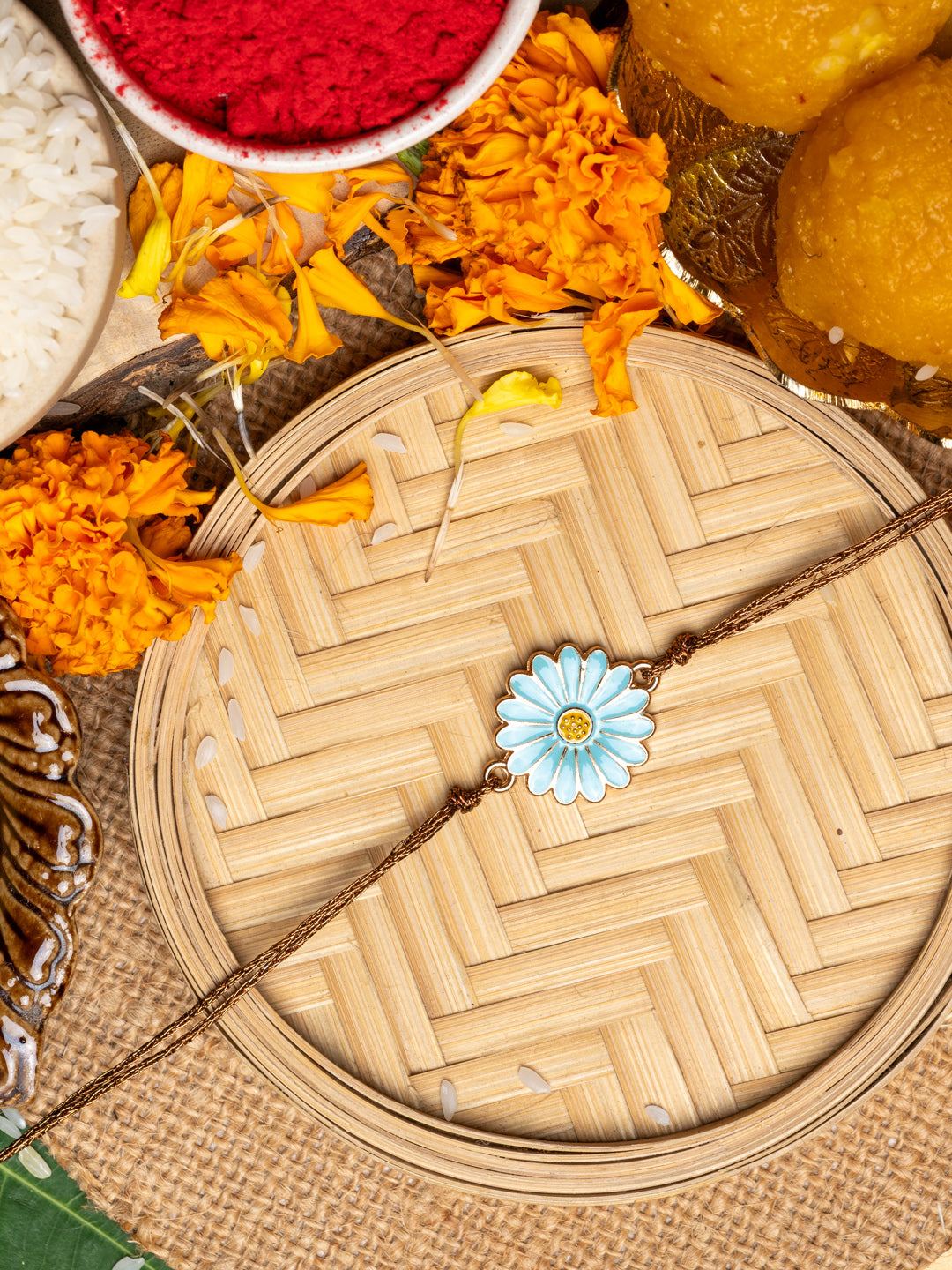 LOOK AND ADORN floral rakhi with blue flower design placed on woven bamboo tray with marigold flowers and traditional sweets