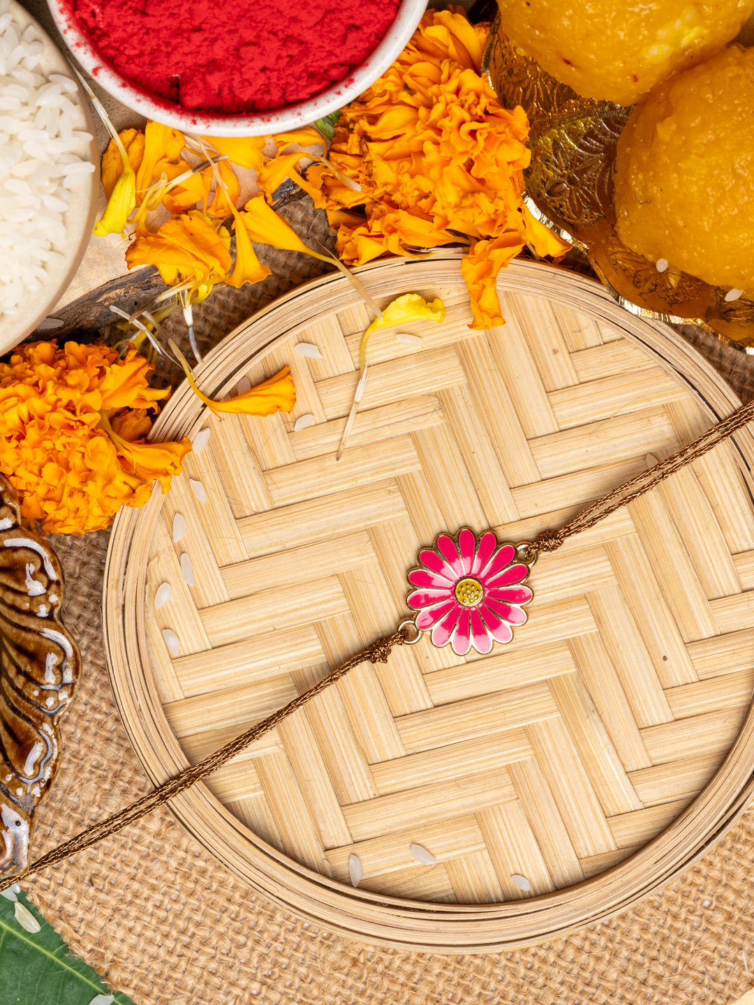 LOOK AND ADORN floral rakhi with pink flower design on woven tray surrounded by marigold flowers and traditional items