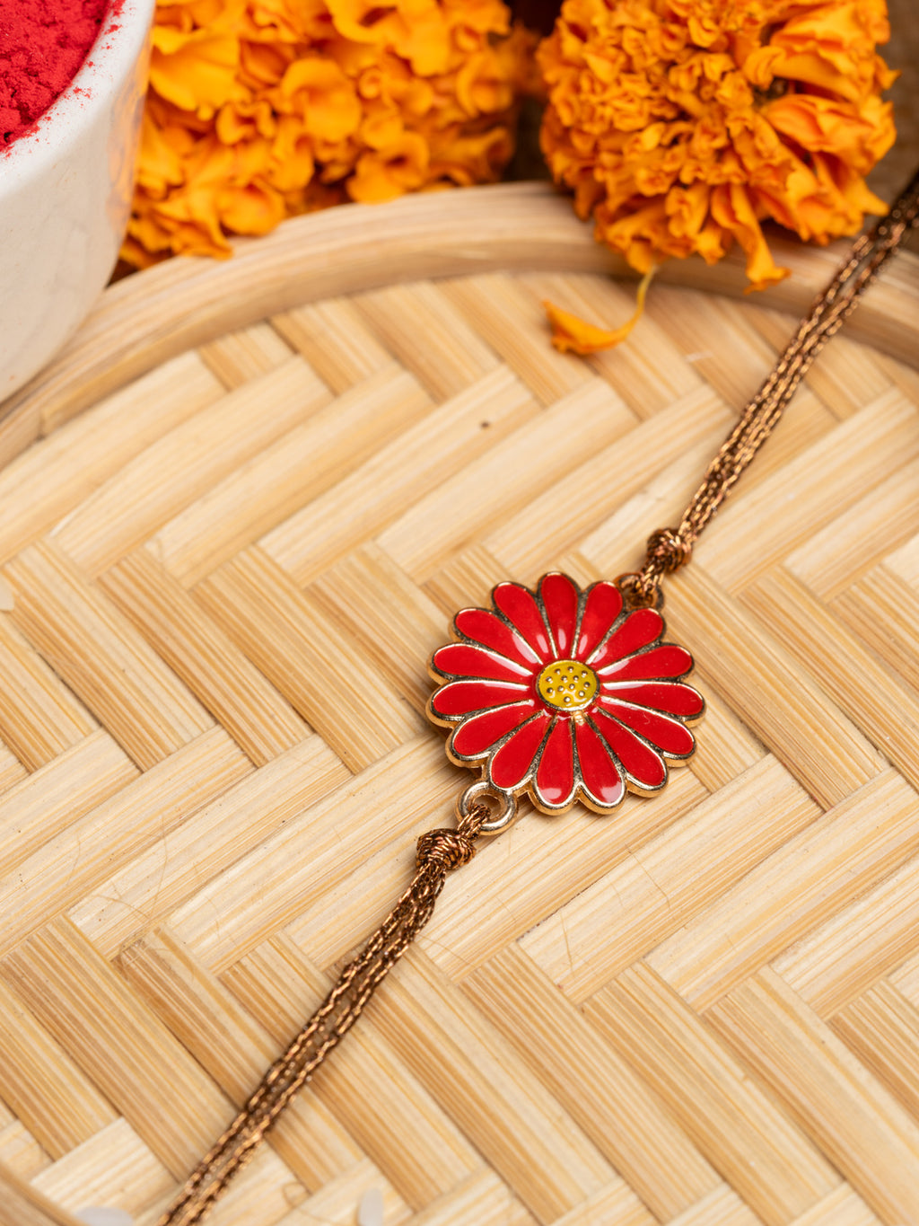 Red floral rakhi with gold chain placed on a woven bamboo mat with marigold flowers in background