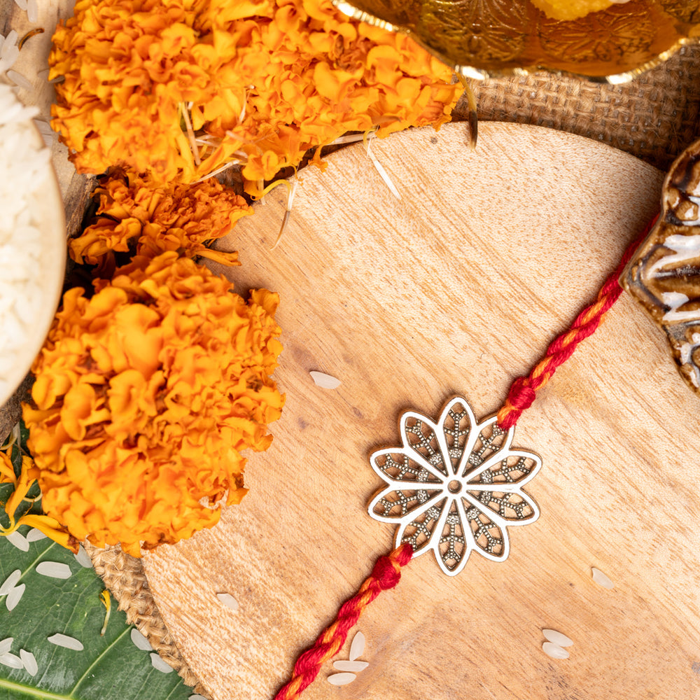 LOOK AND ADORN floral rakhi with silver flower design and red thread on wooden surface with marigold flowers