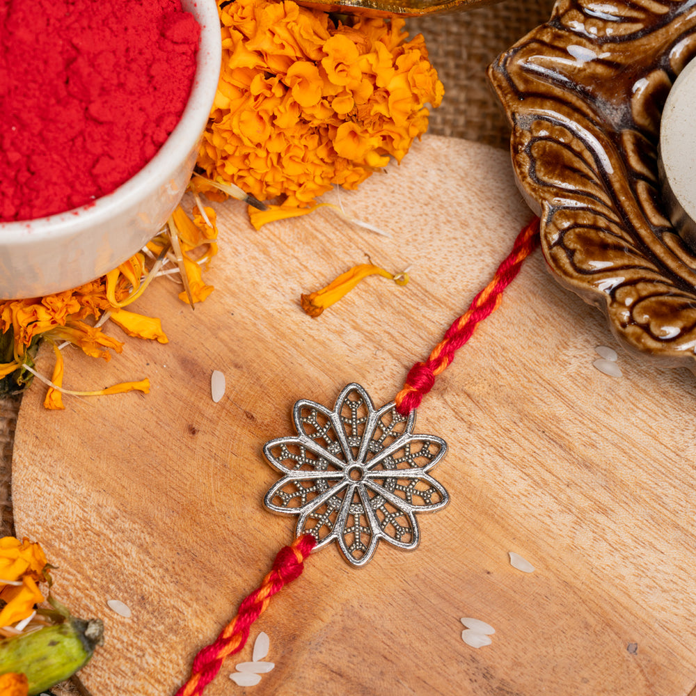 Floral rakhi with silver flower design and red thread placed on wooden surface with marigold and traditional items