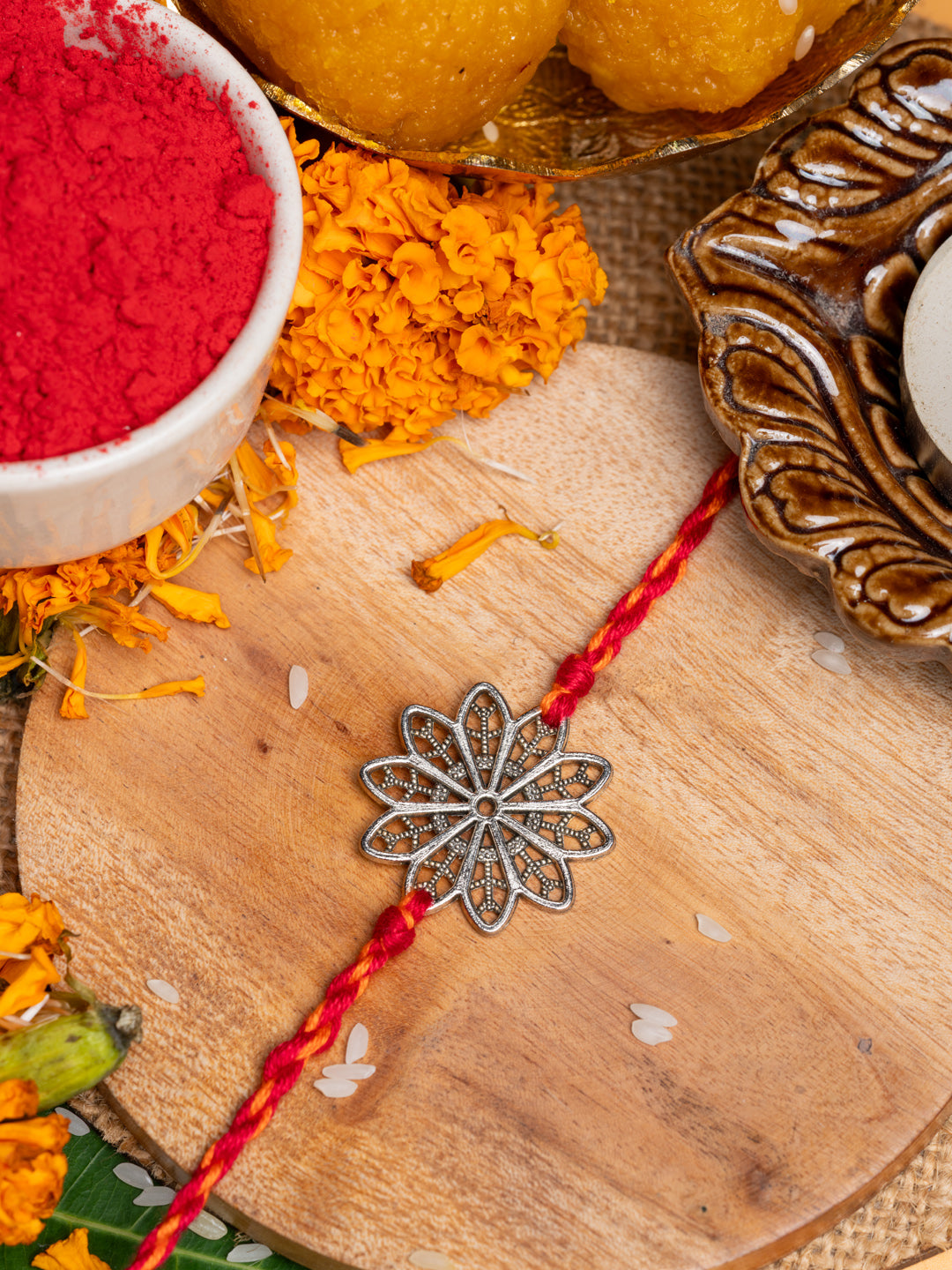 Floral rakhi with silver flower design and red thread placed on wooden surface with marigold and traditional items