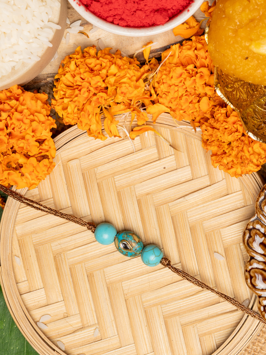 Kundan rakhi with turquoise beads on a woven bamboo tray surrounded by marigold flowers and traditional pooja items