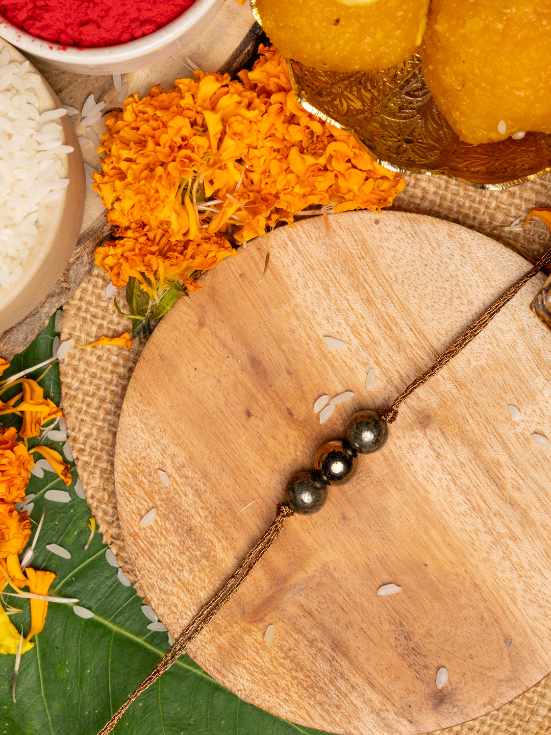 Pyrite rakhi with three metallic beads on golden thread placed on wooden board with marigold flowers and traditional pooja items