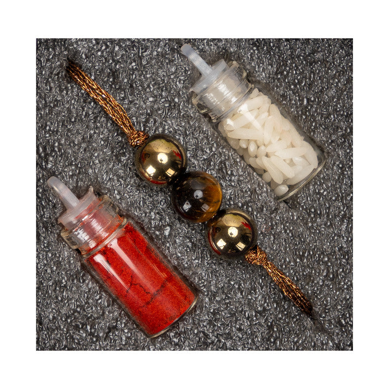 Tiger eye rakhi with gold beads and included red and white powder containers on textured surface