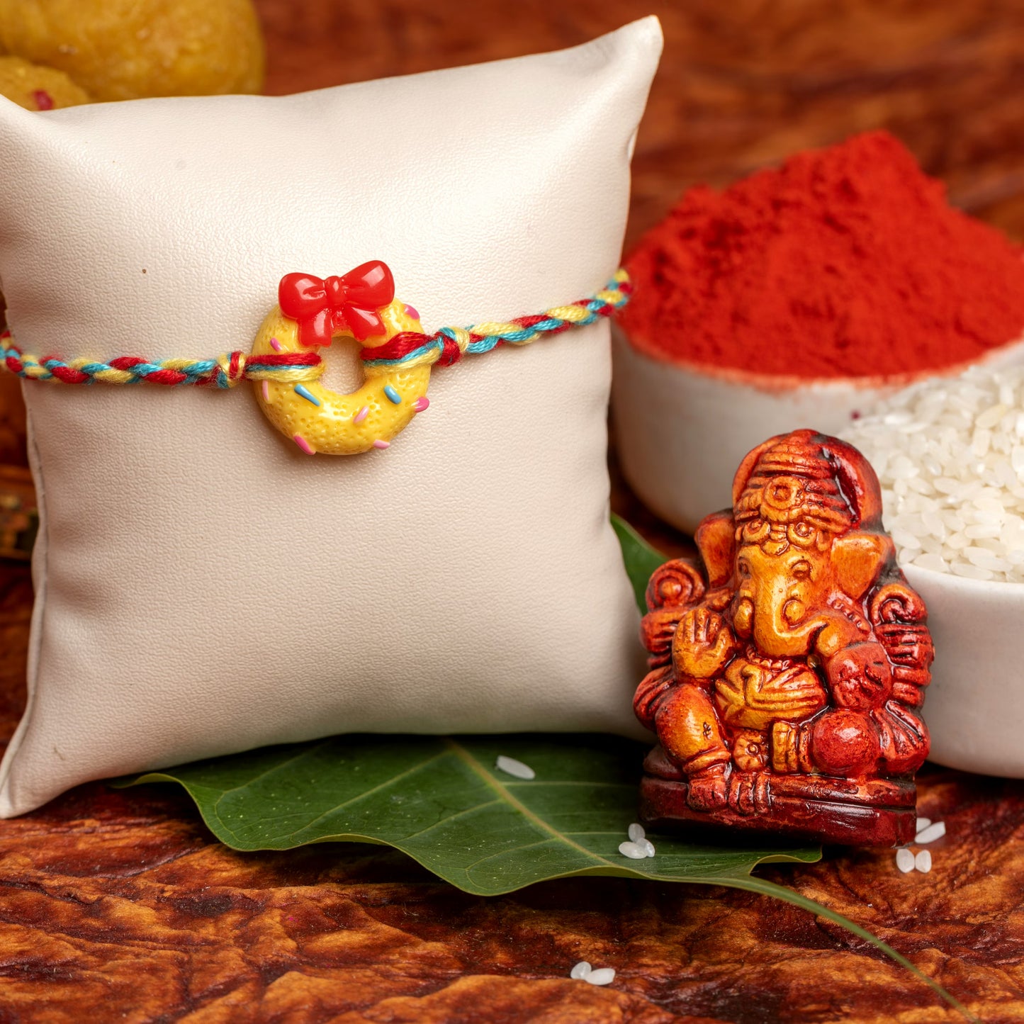 Colorful rakhi with a donut charm and red bow displayed on a white cushion alongside a Ganesha idol and traditional items